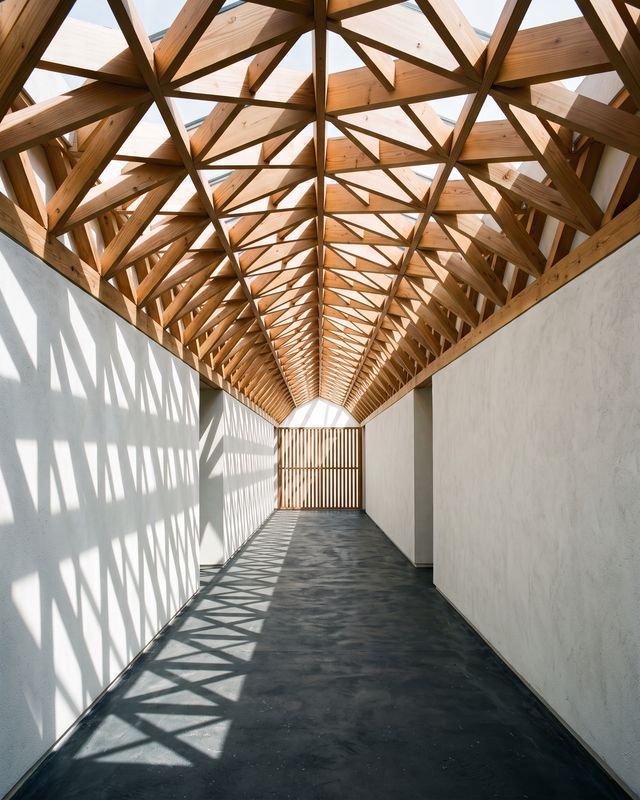 Couloir blanc sous une charpente en bois triangulée, ombres géométriques au sol